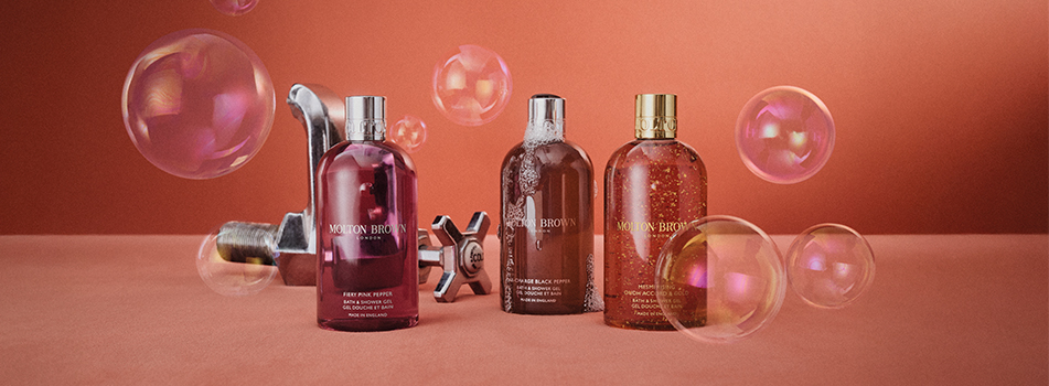 Werbung für Molton Brown Duschgel. Drei Flaschen in verschiedenen Farbtönen – Pink, Braun mit Schaum und Bernstein mit Goldglanz – stehen nebeneinander auf einer orangeroten Fläche. Im Hintergrund befinden sich metallene Armaturen, im Vordergrund schweben schimmernde Seifenblasen. Die Szene wirkt verspielt, luxuriös und frisch.