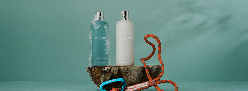 Zwei Flaschen Haarpflegeprodukte von Molton Brown – links eine schäumende, türkisfarbene Shampoo-Flasche, rechts eine weiße Conditioner-Flasche – stehen auf einem Stück Baumrinde vor mintgrünem Hintergrund. Davor liegt ein orangefarbenes Kletterseil mit blauem Karabiner. Die Szene vermittelt Frische, Pflege und Abenteuerlust.