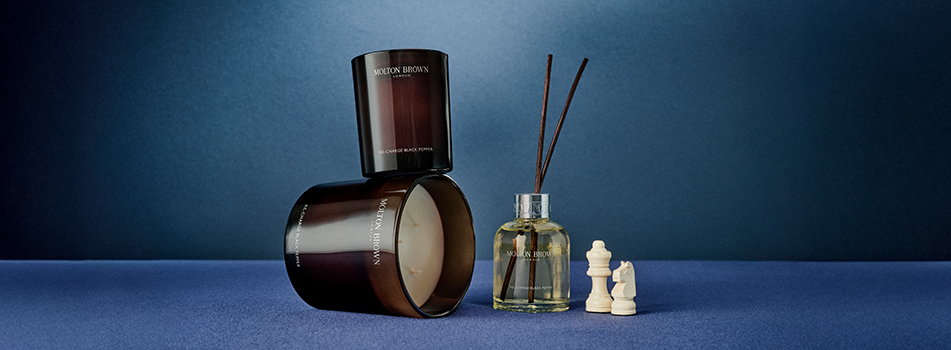 Zwei Duftkerzen von Molton Brown in dunkelbraunen Gläsern, eine stehend, eine liegend mit sichtbarem Wachs, daneben ein Raumduft-Diffuser mit schwarzen Stäbchen. Rechts im Bild stehen drei weiße Schachfiguren – König, Dame und Springer – auf einer blauen Fläche vor einem dunkelblauen Hintergrund. Die Szene wirkt edel, ruhig und stilvoll.